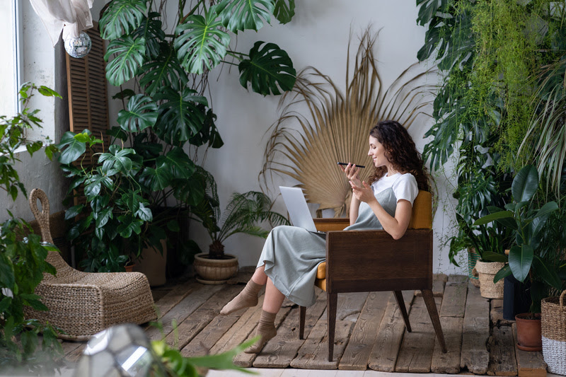 Positive female with eyes closed sending voicemail, using laptop, sitting on chair in green house with evergreen exotic monstera plants. Young woman entrepreneur work on laptop, sitting in homegarden.